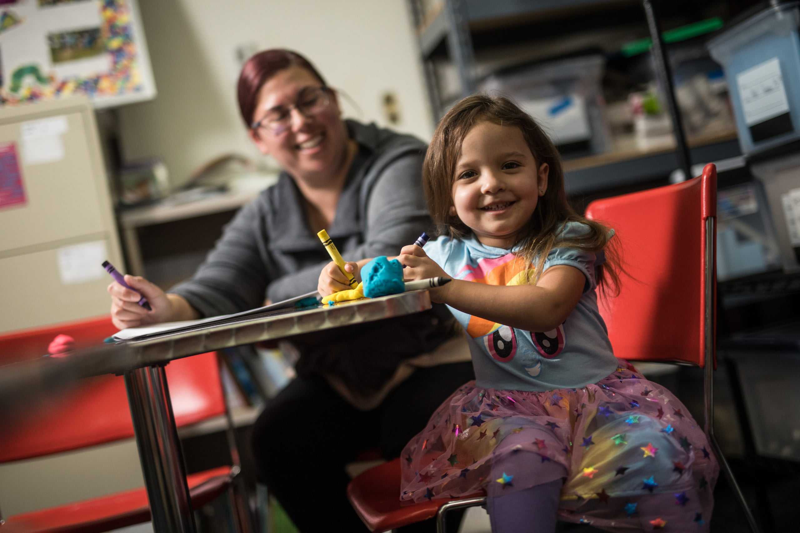 Smiling child with crayons, supervised by a woman